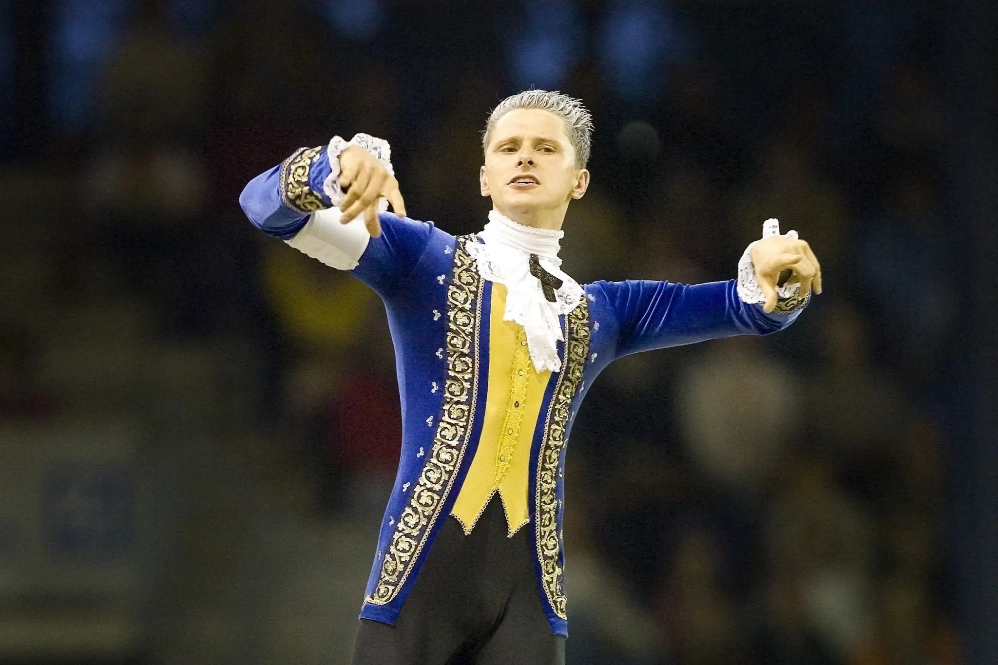 A vaulter stands in a blue and yellow baroque costume on the back of a horse. The costume is inspired by the composer Mozart. The scene takes place in an illuminated arena in front of a blurred audience.