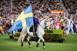 A rider on a white horse carries a large Swedish flag through a packed stadium; in the background are more flags, spectator stands and colourful decorations during the march-in of the nations.
