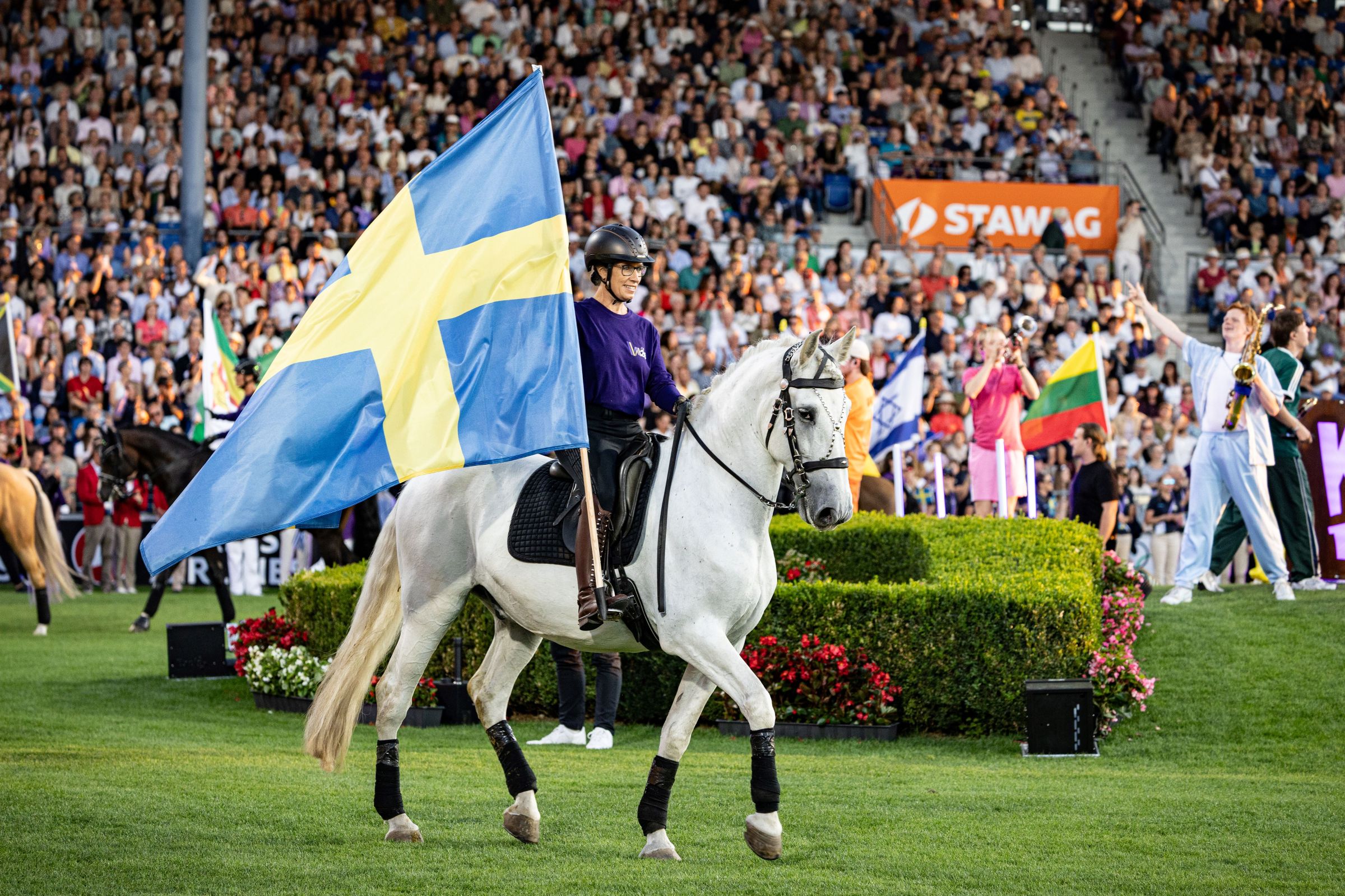 A rider on a white horse carries a large Swedish flag through a packed stadium; in the background are more flags, spectator stands and colourful decorations during the march-in of the nations.