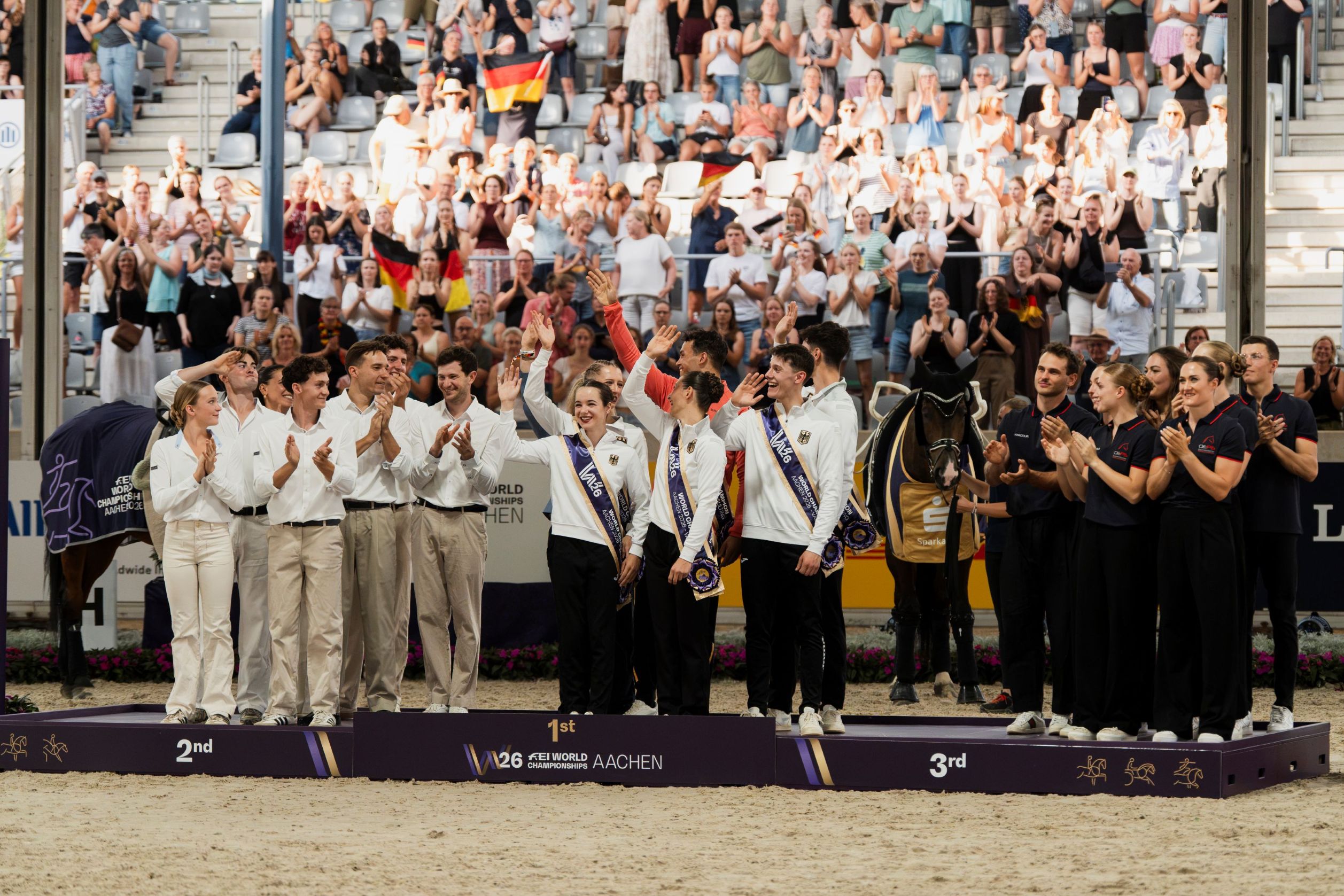 Vaulting medal ceremony: A group of young vaulters in black-and-white team kit bearing the German emblem and sashes stand jubilantly on the podium, flanked on the left and right by teams of coaches and support staff; in the background, an applauding crowd waves German flags, and a horse, fully kitted out and ready for vaulting, stands nearby.