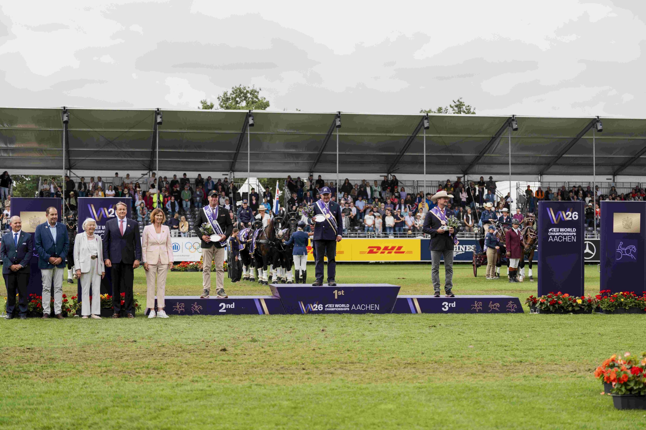 Siegerehrung im Gespannfahren bei den FEI World Championships Aachen 2026: Drei ausgezeichnete Fahrer stehen mit ihren Pferdegespannen auf dem Podium, umgeben von Offiziellen und vor voll besetzten Zuschauertribünen.