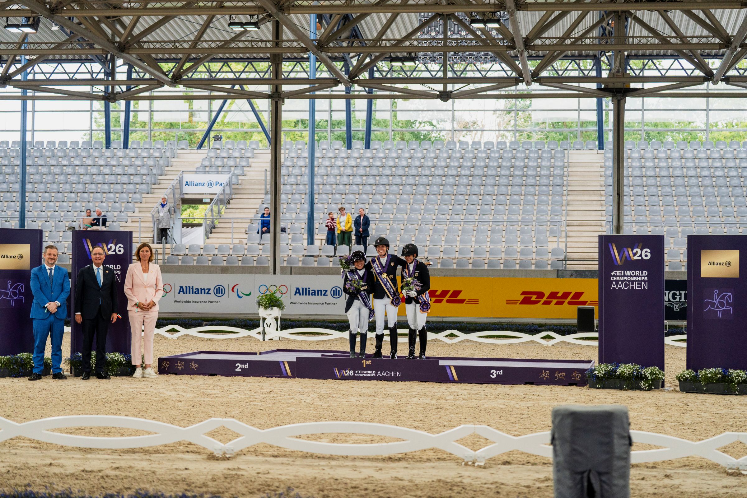 Siegerehrung in der Para-Dressur in Grade IV bei den FEI World Championships Aachen 2026: Drei ausgezeichnete Reiterinnen und Reiter stehen mit Medaillen und Blumen auf dem Podium, begleitet von Ehrengästen in einem Reitstadion.