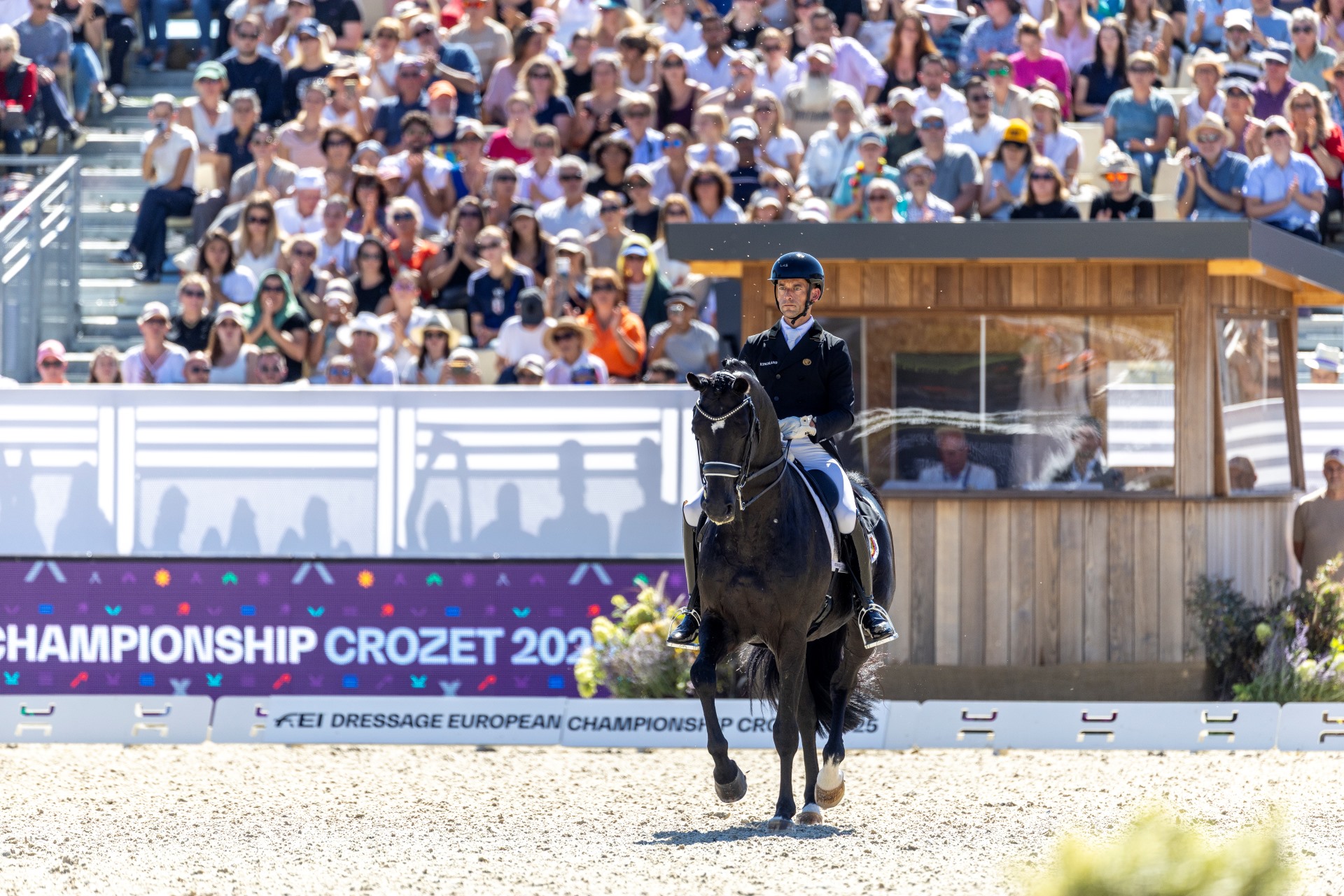 A dressage rider on a black horse rides around the competition arena in front of a packed grandstand; in the background is a large banner reading “CHAMPIONSHIP CROZET” from the FEI Dressage European Championship.