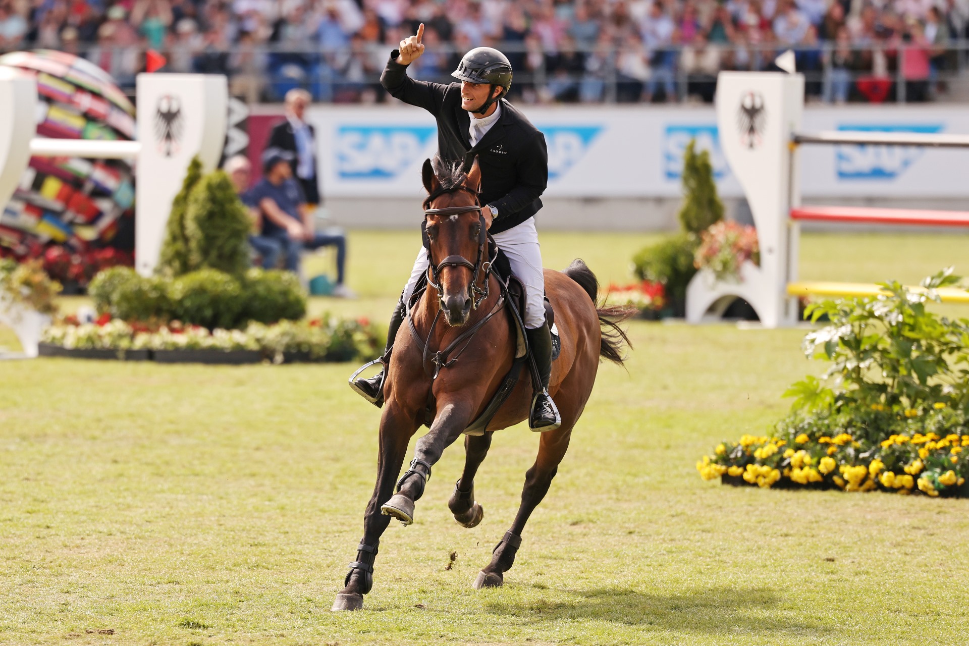 Show jumper on a brown horse gallops victoriously across a grass arena, raising his right arm with one finger pointed upward; in the background are fences with a federal eagle emblem, sponsor boards, and packed spectator stands.