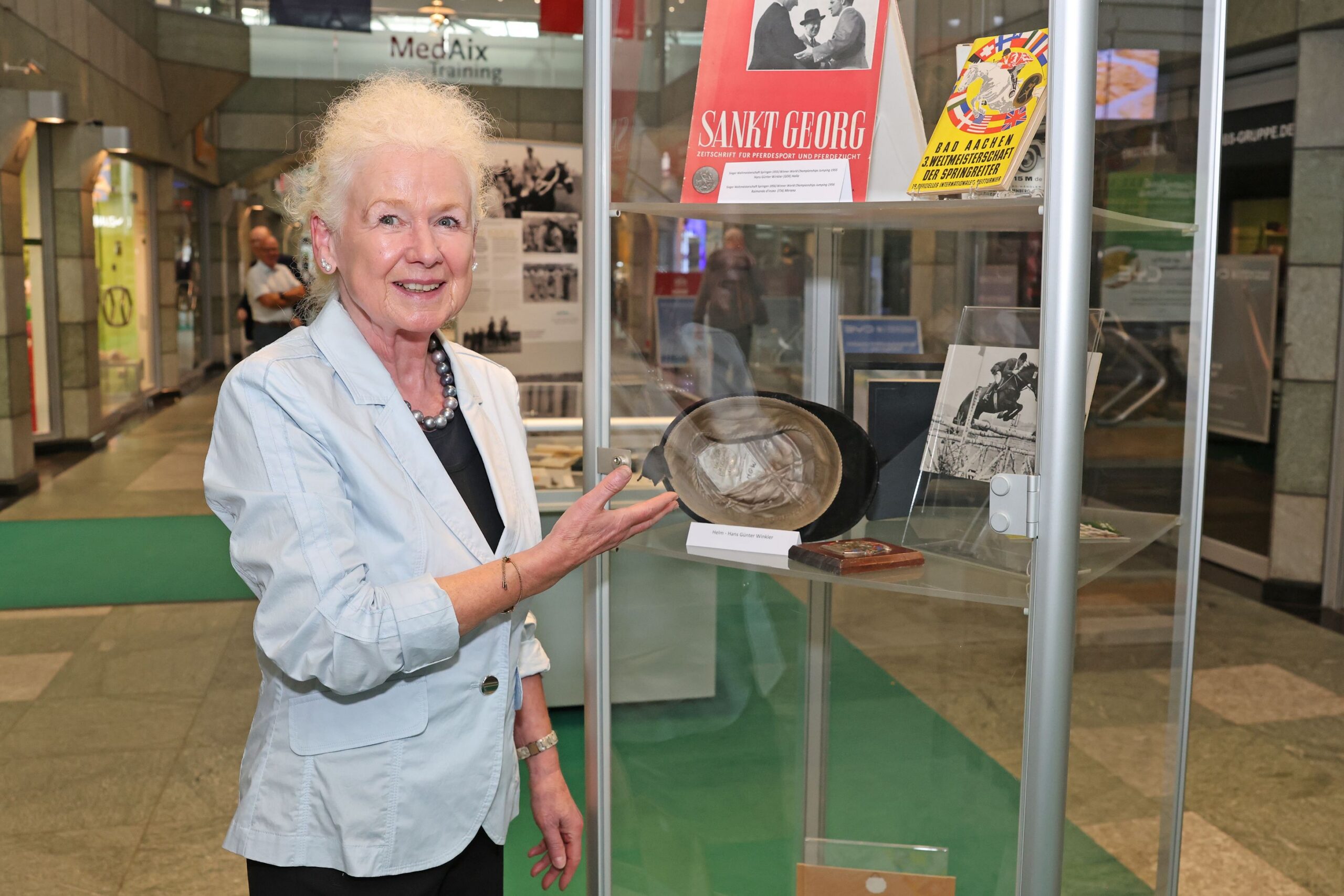 A woman in a light blue blazer is standing in a shopping arcade in front of a glass display case at an equestrian exhibition and smiling at the camera; with her right hand, she is pointing at an old riding helmet on display, next to which lie historical magazines and black-and-white photographs from the world of equestrian sport.