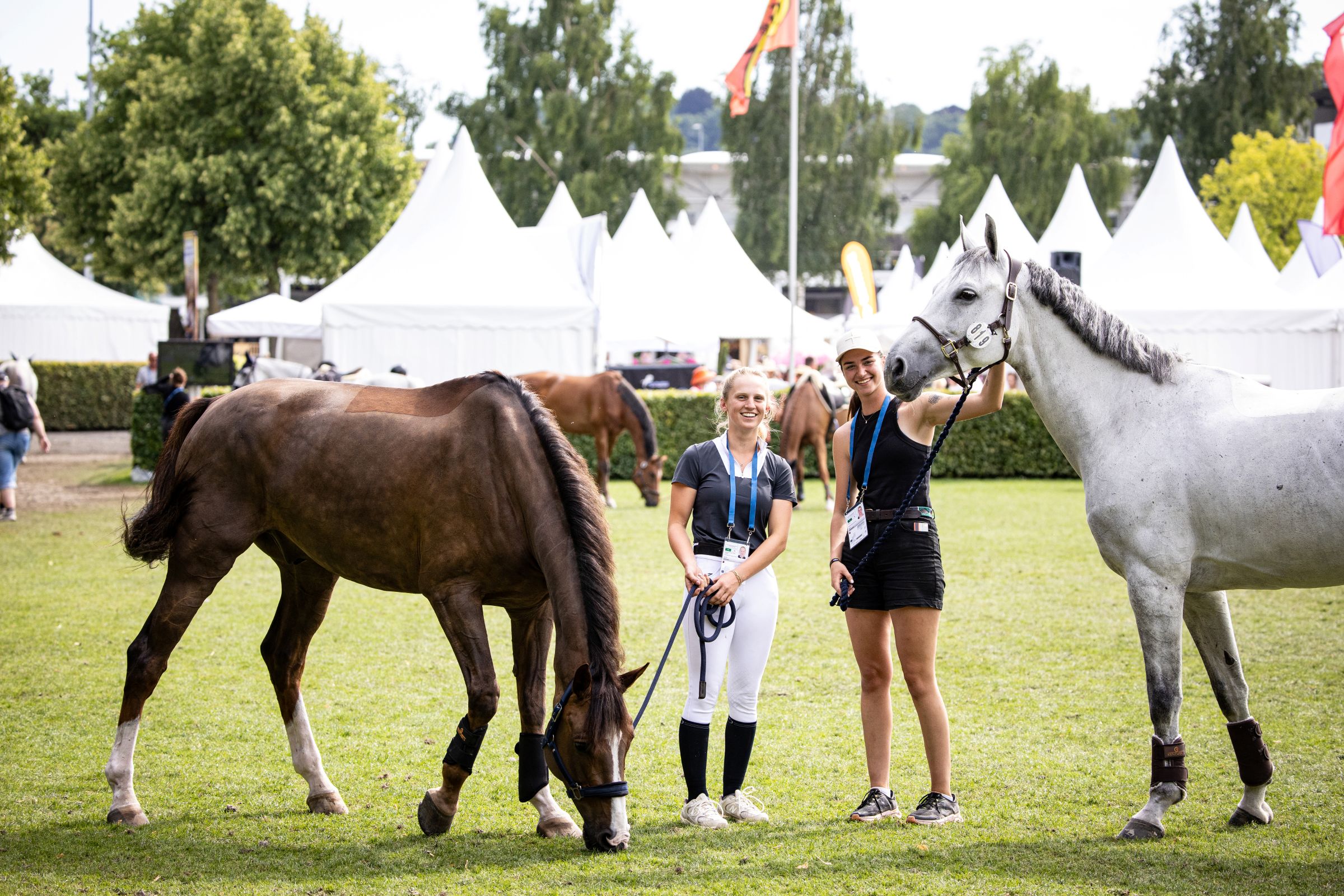 Two young women are standing in a meadow, laughing, between a brown horse and a grey horse that are grazing; they are holding them by their halters; in the background, white tents, trees and the hustle and bustle of the show can be seen.