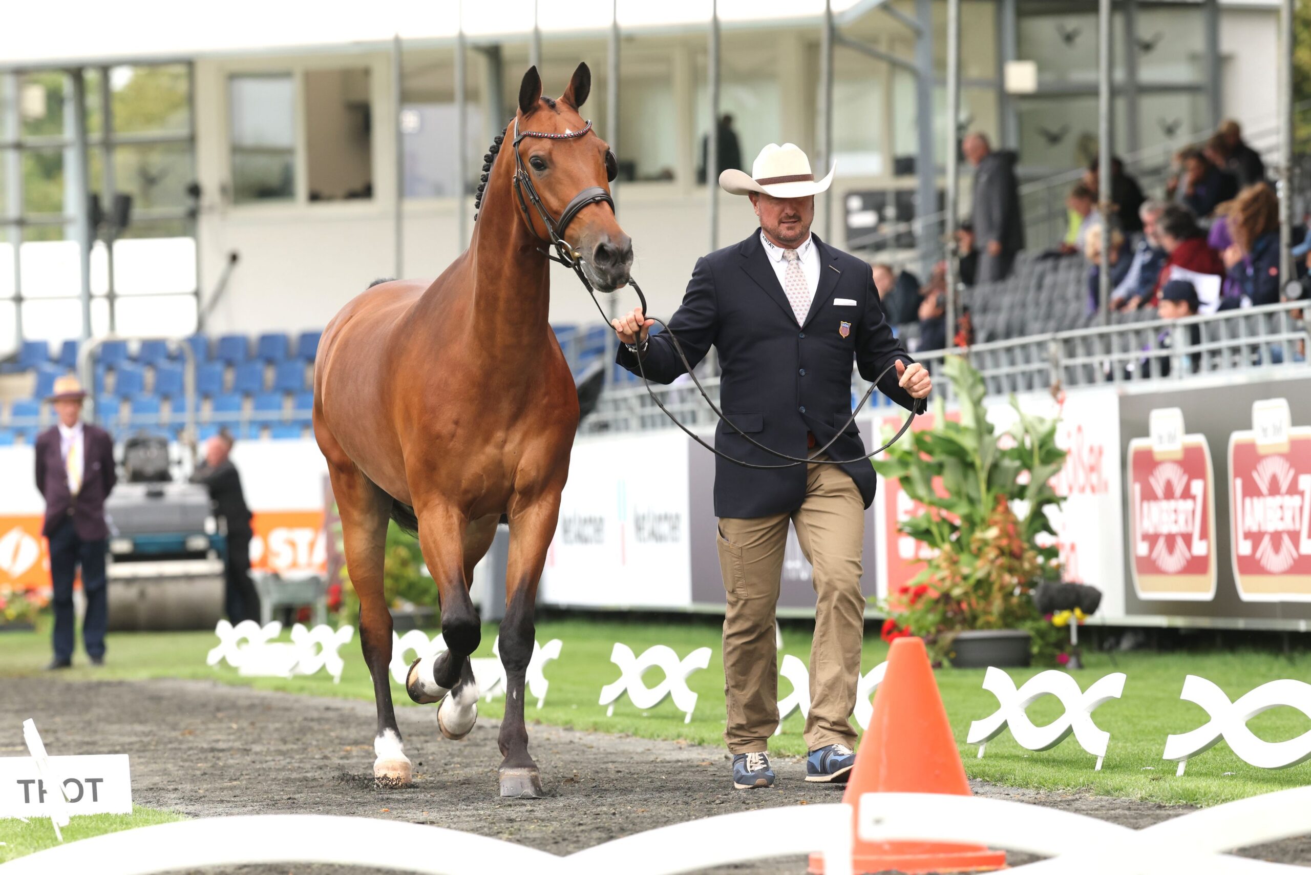 A brown horse is being led through a riding arena by a man in a dark jacket and a white cowboy hat; in the background, spectators are seated in the stands.