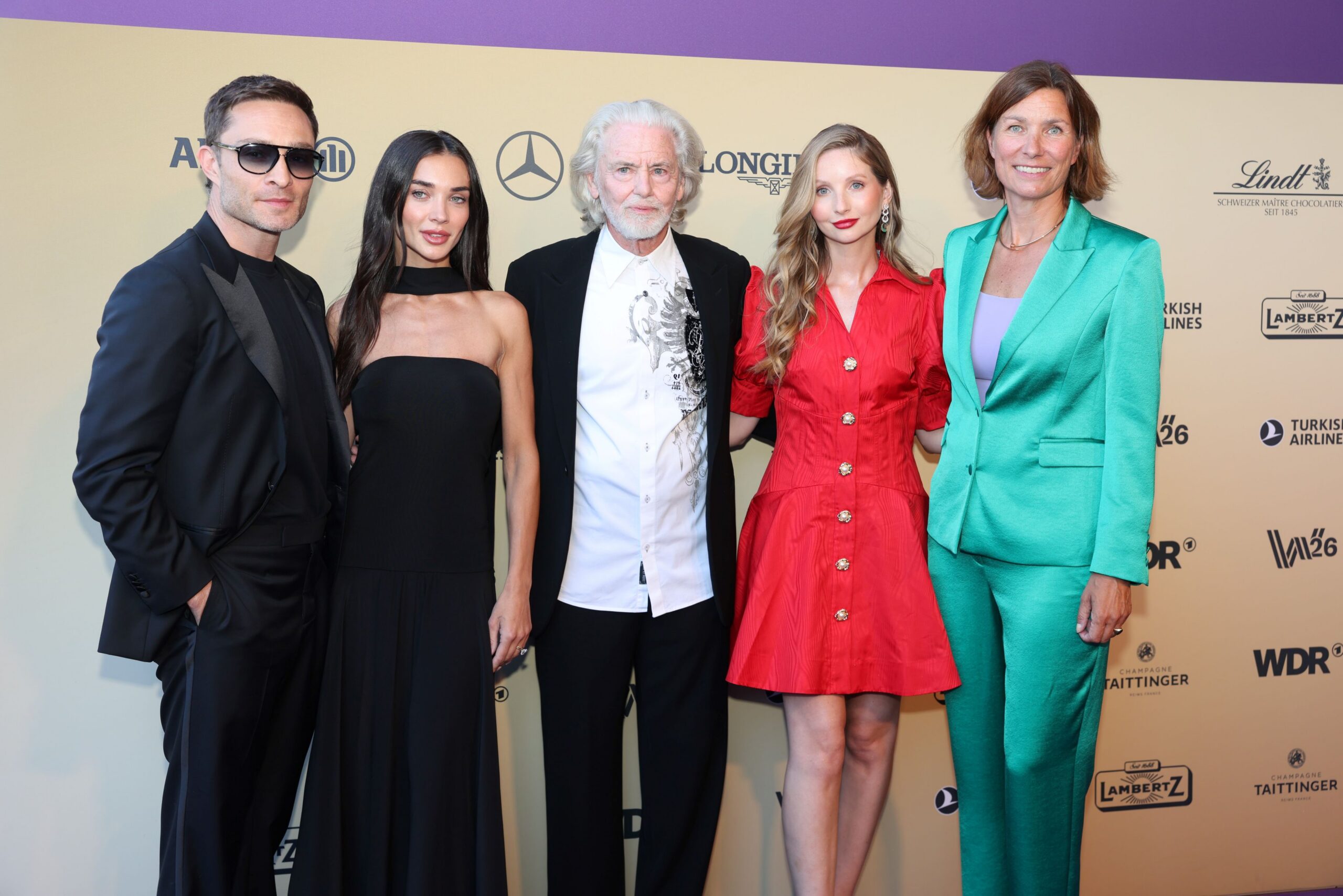 Eine Gruppe von fünf Personen posiert vor einer goldfarbenen Event Wand mit Logos: von links ein Mann im schwarzen Anzug mit Sonnenbrille, daneben eine Frau im schulterfreien schwarzen Kleid, in der Mitte ein Mann mit weißem Haar in weißem Hemd und dunklem Jackett, neben ihm eine Frau in einem roten Kleid und rechts eine Frau in einem türkisfarbenen Hosenanzug, alle lächelnd in die Kamera.