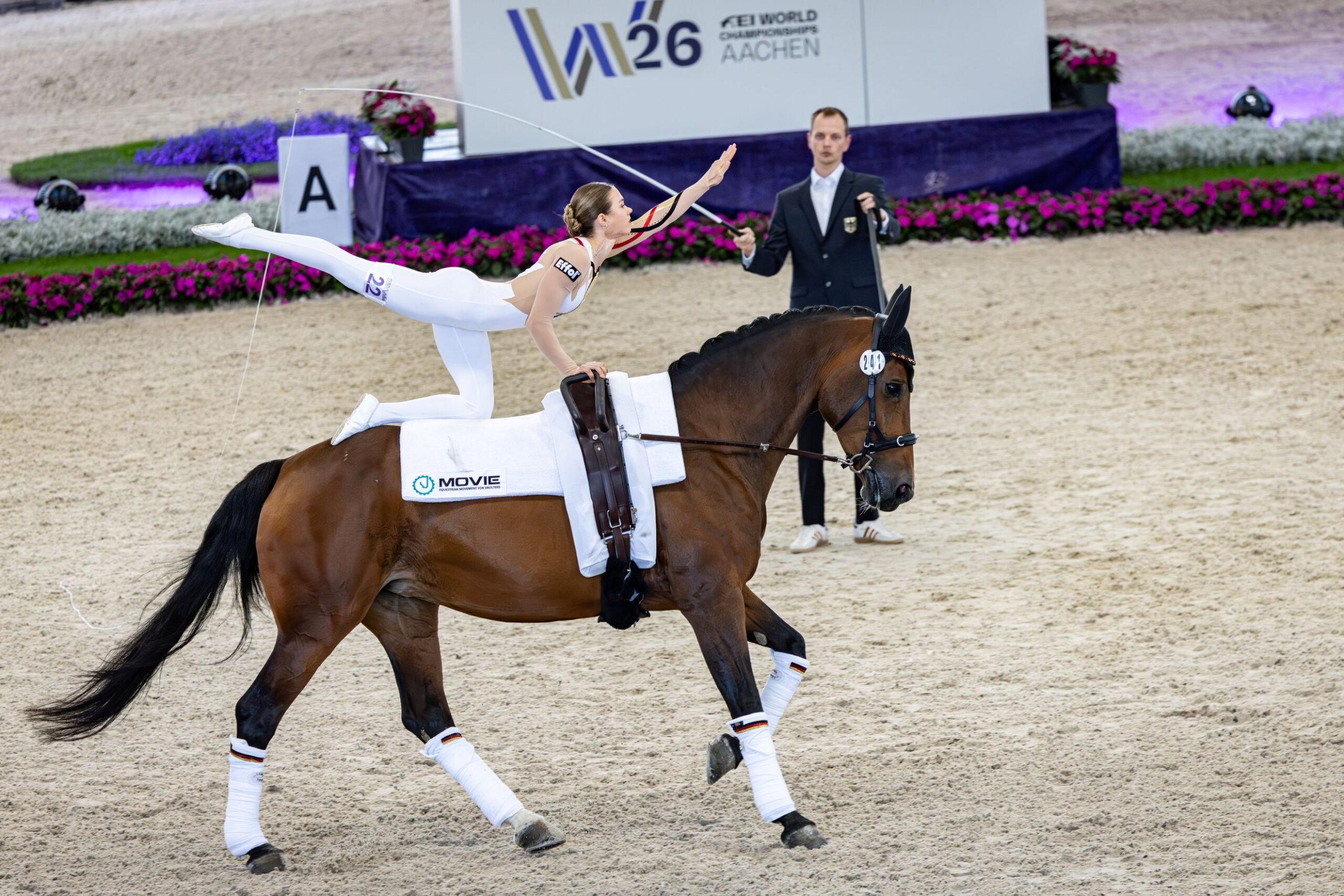 A vaulter in a white leotard is balancing in a high lateral position on a galloping horse, whilst in the background the lunger and the flower-decorated perimeter advertising featuring the logo of the FEI Vaulting World Championship Aachen 2026 can be seen.