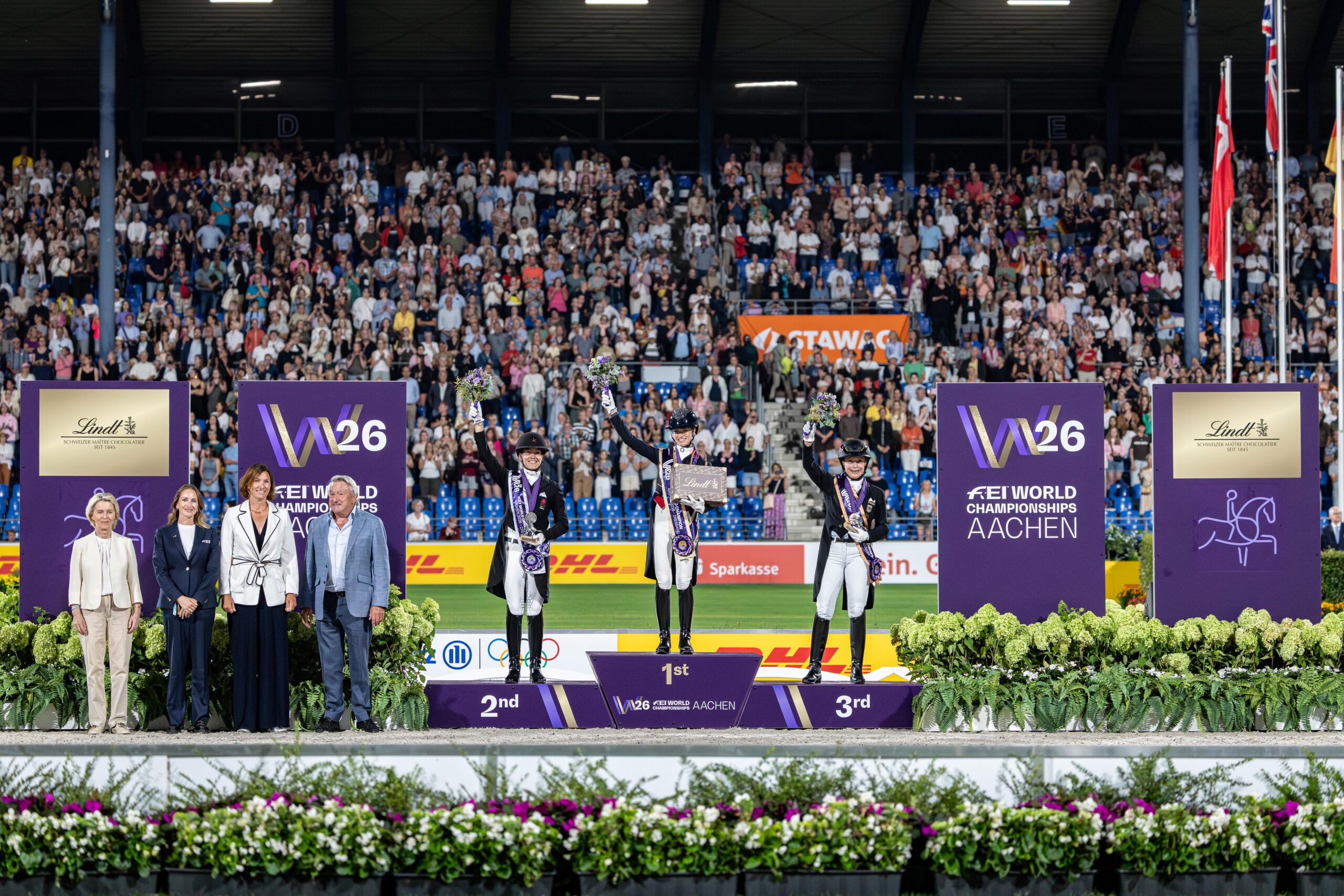 Dressage prize-giving ceremony at the FEI World Championships Aachen 2026: Three riders in tailcoats, riding helmets and sashes stand jubilantly on the podium with bouquets of flowers in front of a packed, applauding grandstand, flanked by officials and large purple competition backdrops featuring W26 and FEI branding.