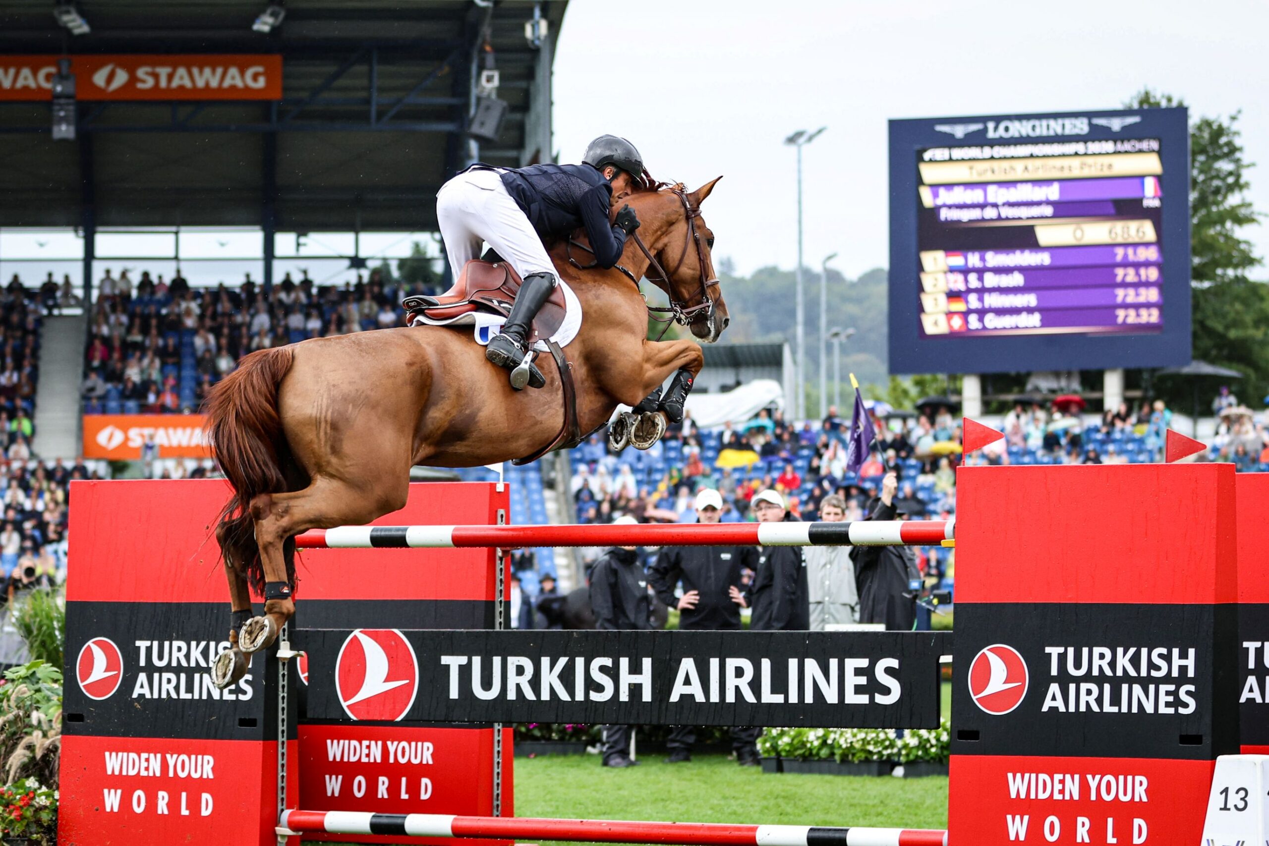 A rider jumps over a red-and-white obstacle bearing the words ‘Turkish Airlines’ with his bay horse in the equestrian stadium. In the background, spectators’ stands and a scoreboard can be seen.
