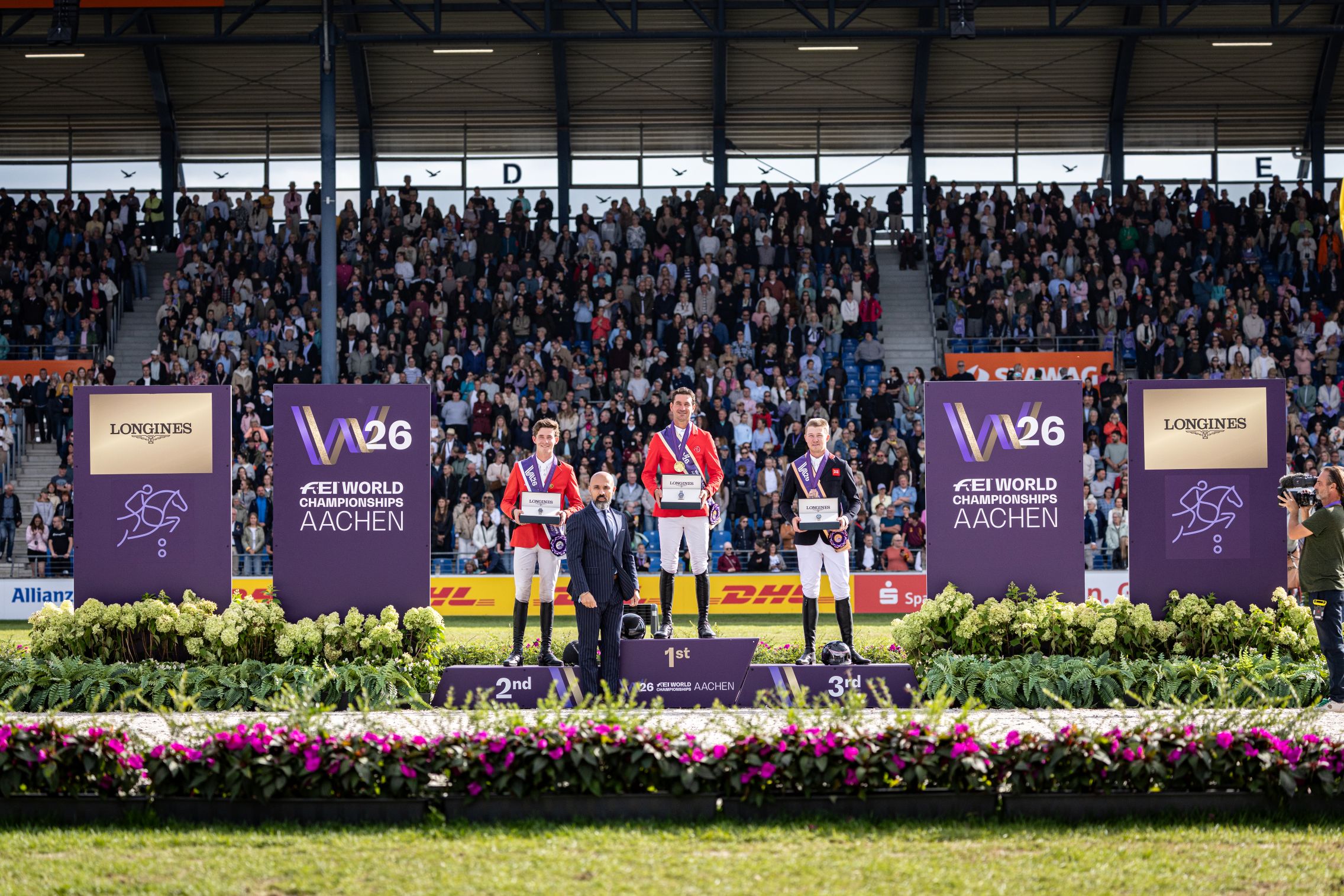 Drei Reiter stehen bei der Siegerehrung der FEI-Weltmeisterschaften Aachen 2026 auf dem Podium: Der Sieger in der Mitte auf Platz 1, flankiert vom Zweit- und Drittplatzierten. Dahinter applaudiert das Publikum; Blumen, Sponsorenlogos und Tafeln mit der Aufschrift „FEI World Championships Aachen“ rahmen die Szene ein.