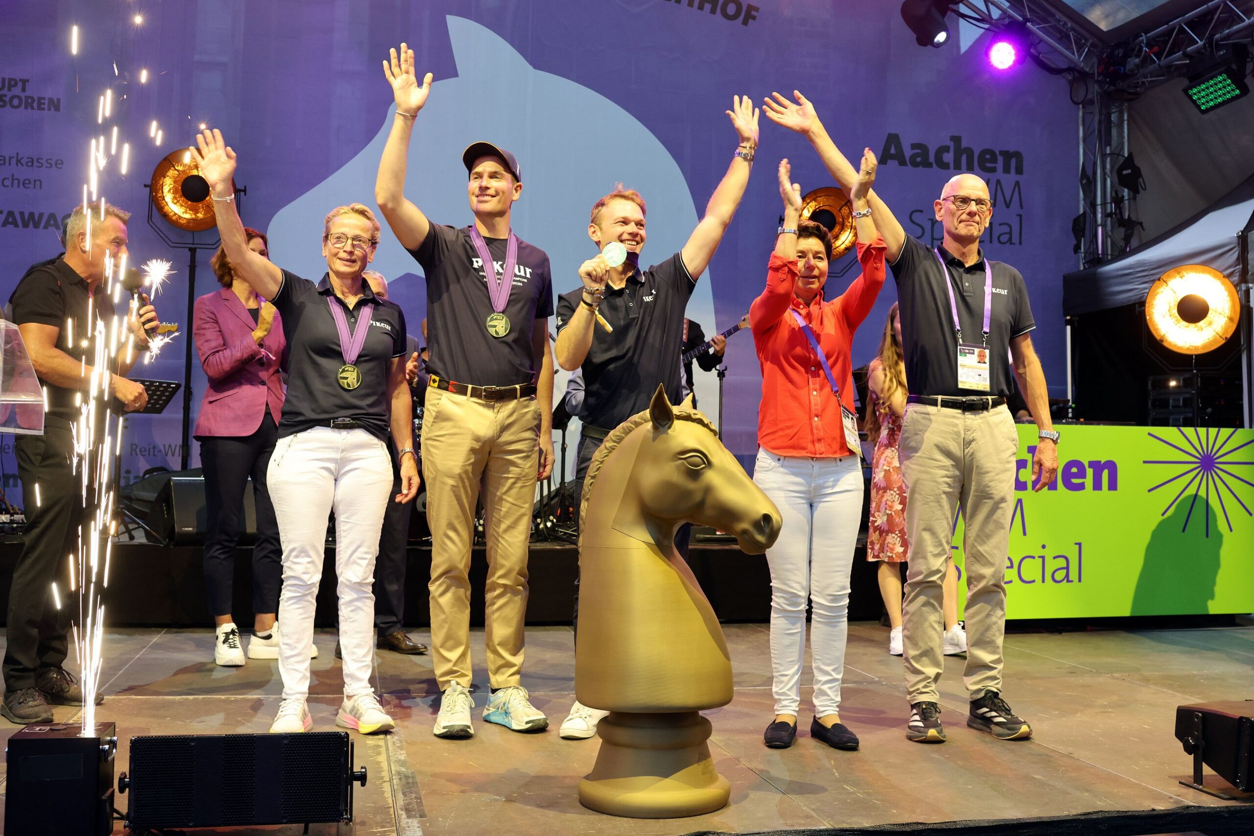 A group of five honourees on stage at the Aachen WM Special, all wearing medals round their necks, standing next to a large golden horse’s head trophy, laughing and waving; on the left, stage fountains are spraying water, whilst in the background there are musicians, spotlights and a banner bearing the word ‘Aachen’.