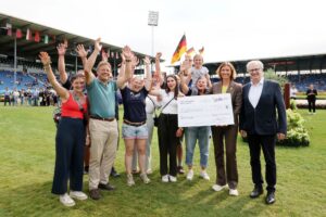 A group of cheerful people on the pitch at the Main Stadium, cheering and throwing their arms in the air as they hold aloft a large donation cheque for 5,000 euros; in the background, the spectator stands can be seen, decorated with national flags, whilst several German flags are visible on the pitch.