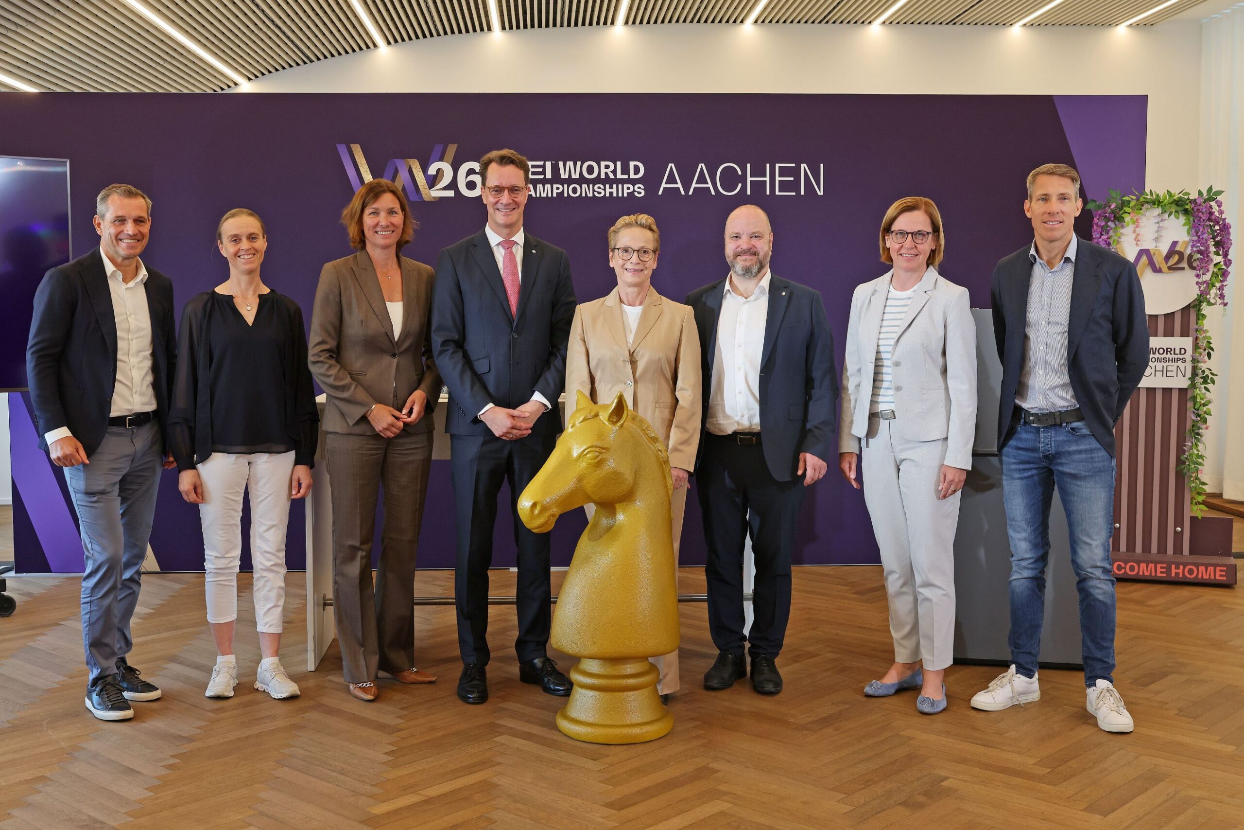 Eight people standing in a row in front of a purple wall bearing the text ‘W26 FEI World Championships Aachen’; a large, golden sculpture of a horse’s head is stood on a wooden floor in the centre of the group.