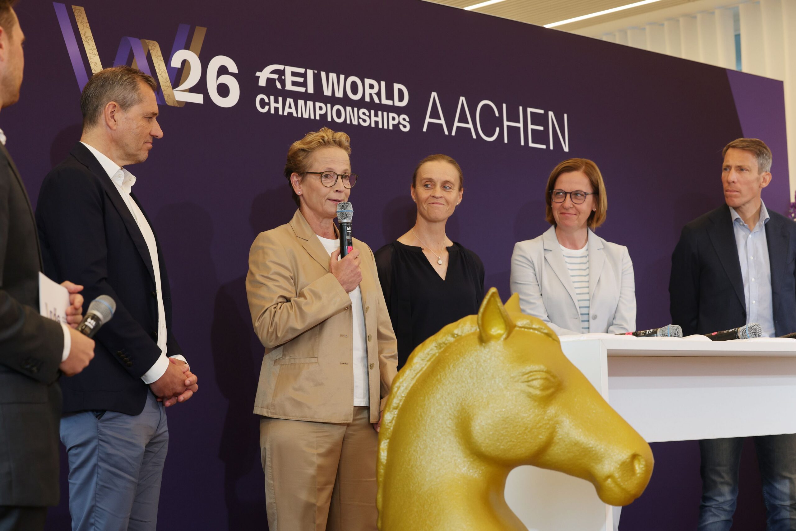 Several people standing in front of a purple wall bearing the text "W26 FEI World Championships Aachen"; a woman in a beige suit is speaking into a microphone, flanked by other participants, while a large golden sculpture of a horse’s head is visible in the foreground.