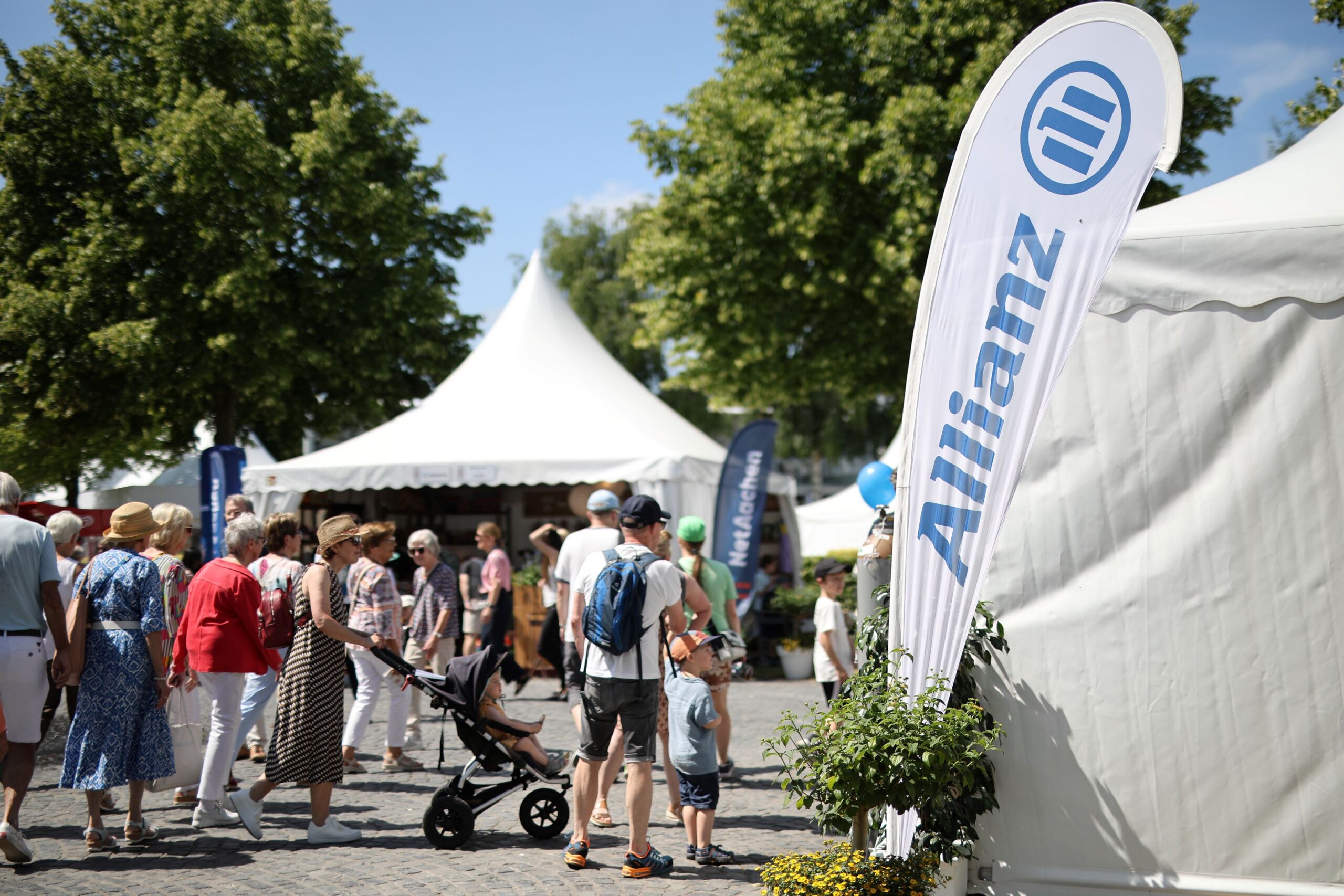 In sunny weather, people are strolling across an outdoor area dotted with white pagoda tents; in the foreground is a child in a pushchair, next to him a man with a backpack, and to the right a white beach flag bearing the blue Allianz logo rises up into the air.