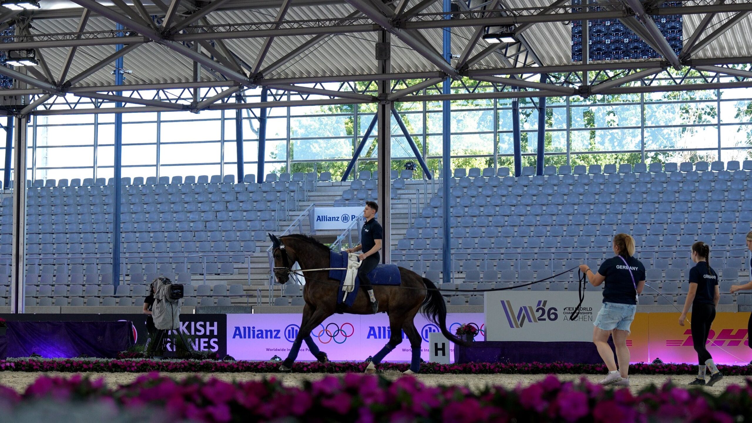 Vaulting training in a brightly lit indoor arena: an athlete sits hands-free in the saddle on a bay horse, which is being led on the lunge by team members wearing ‘Germany’ shirts; in the foreground, pink floral decorations; in the background, empty stands and hoardings bearing the Allianz and FEI World Championships Aachen 2026 logos.