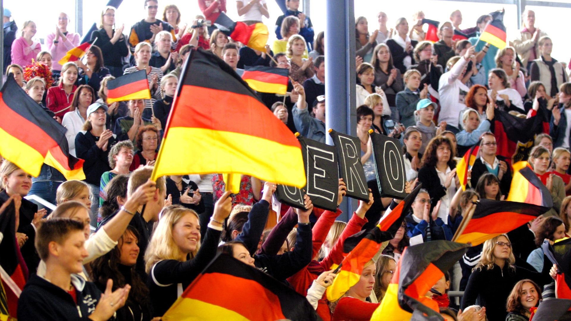 Crowd of spectators in the stands enthusiastically waving black-red-gold German flags and clapping, creating a festive atmosphere at an event in an arena.