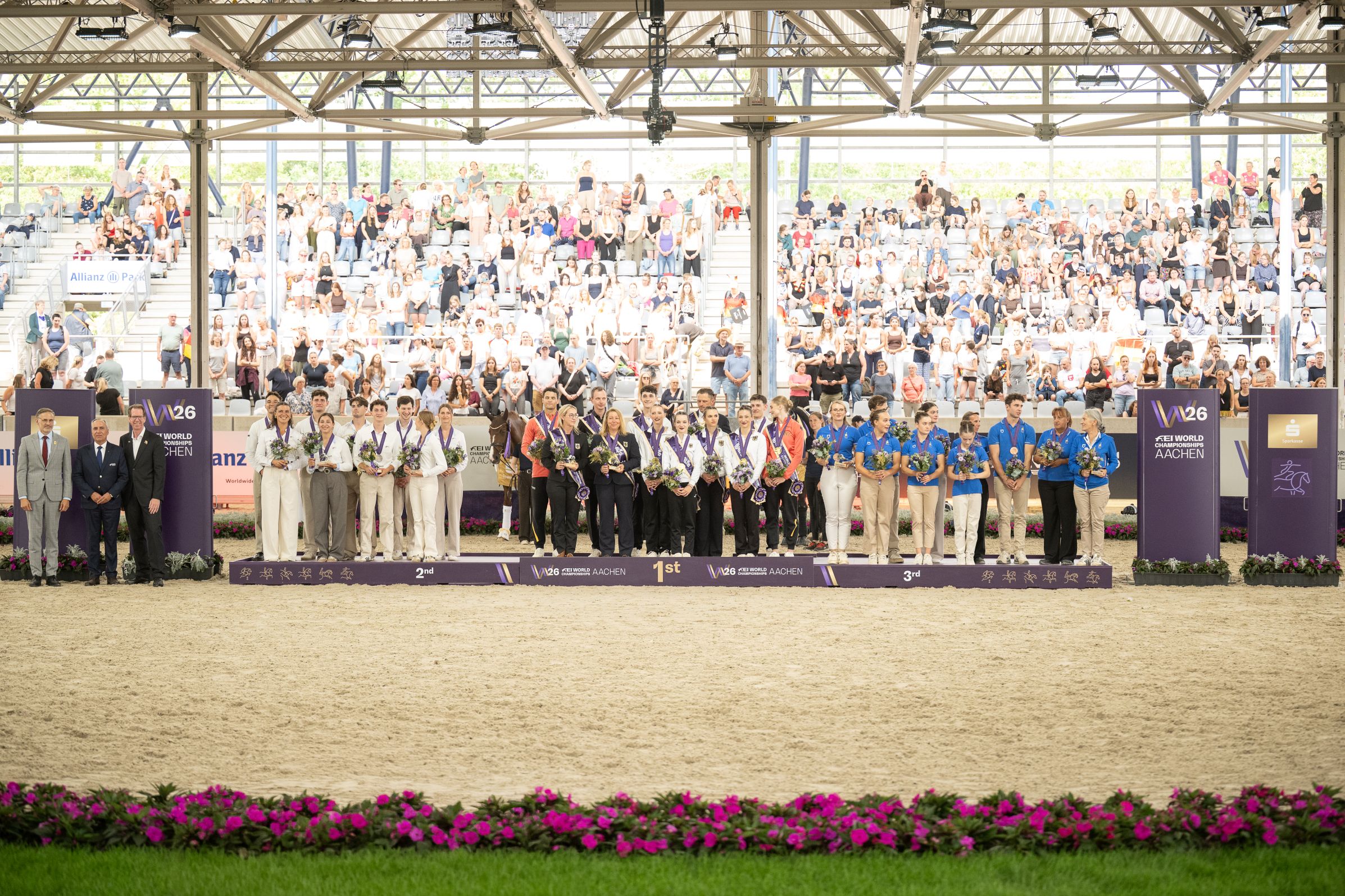 Medal Ceremony at the FEI World Championships in Aachen: Several vaulting teams, dressed in their competition outfits and holding medals and bouquets of flowers, stand on a wide podium bearing the inscriptions ‘2nd’, ‘1st’ and ‘3rd’, flanked on the right and left by officials. In the background, a large, covered circular grandstand filled with numerous spectators completes the picture; at the front, a flowerbed with pink blooms lines the sand arena.