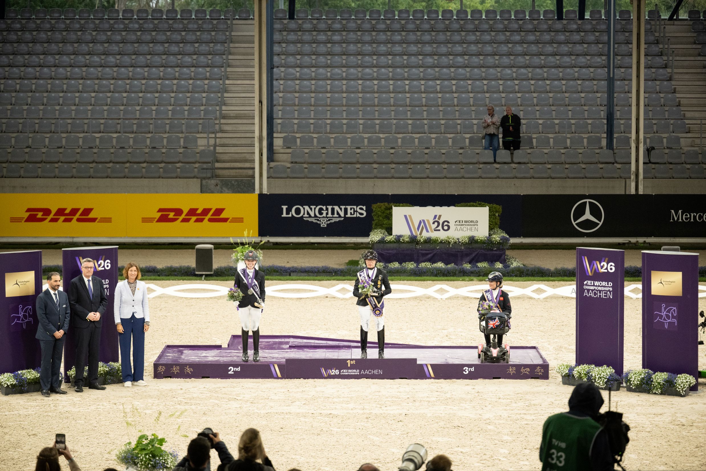 Medal Ceremony at Stadium 3: Three riders stand on the podium in first, second and third place, flanked by four guests of honour. In the background, the stands, sponsors’ logos and the logo of the FEI World Championships Aachen 2026 can be seen.