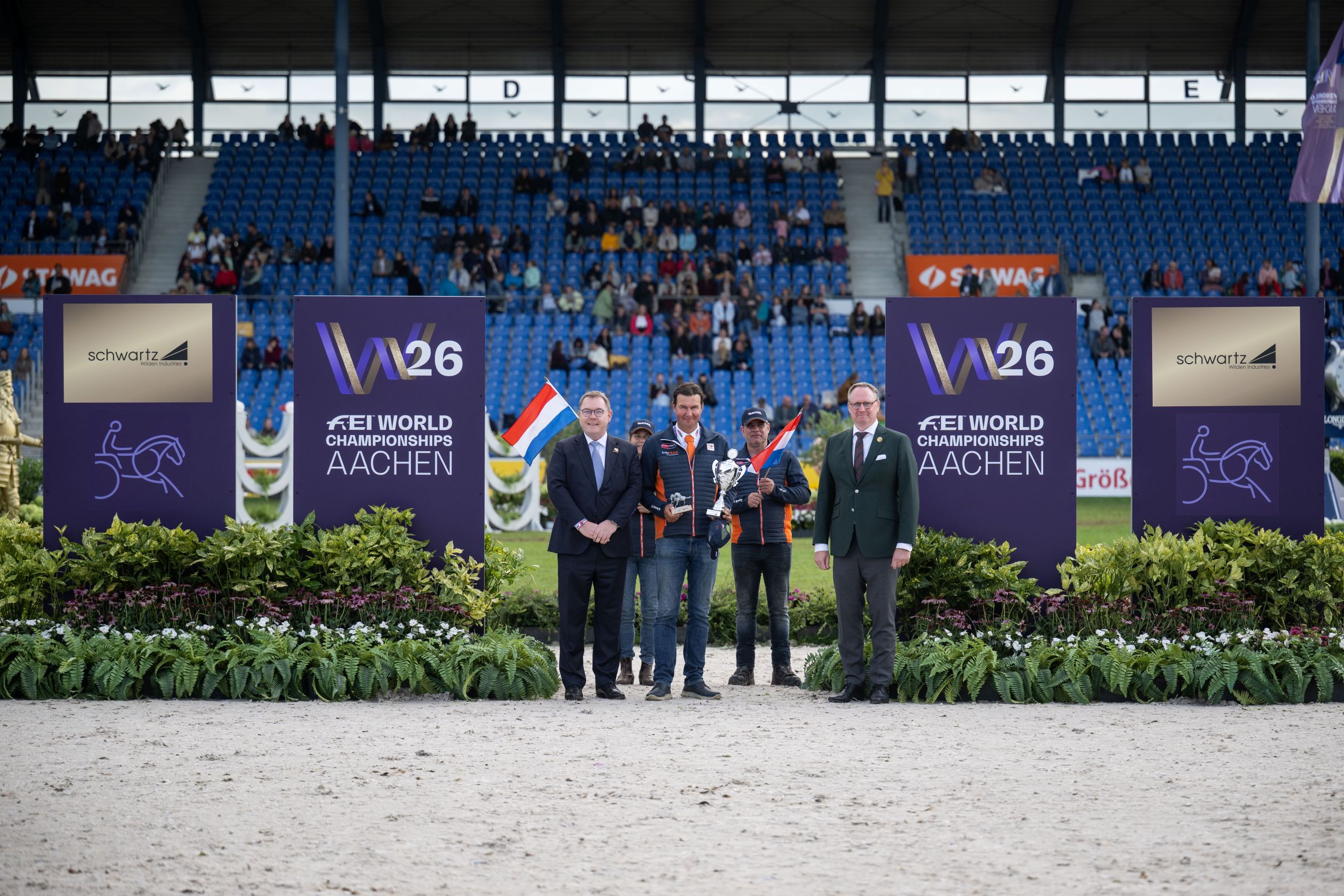 Siegerehrung im Stadion: Fünf Männer stehen neben großen „W26 FEI World Championships Aachen“-Tafeln und „schwartz“-Sponsorboards; einer hält die Trophäe, sein Team hält niederländische Flaggen, im Hintergrund sind die Tribünen zu sehen.