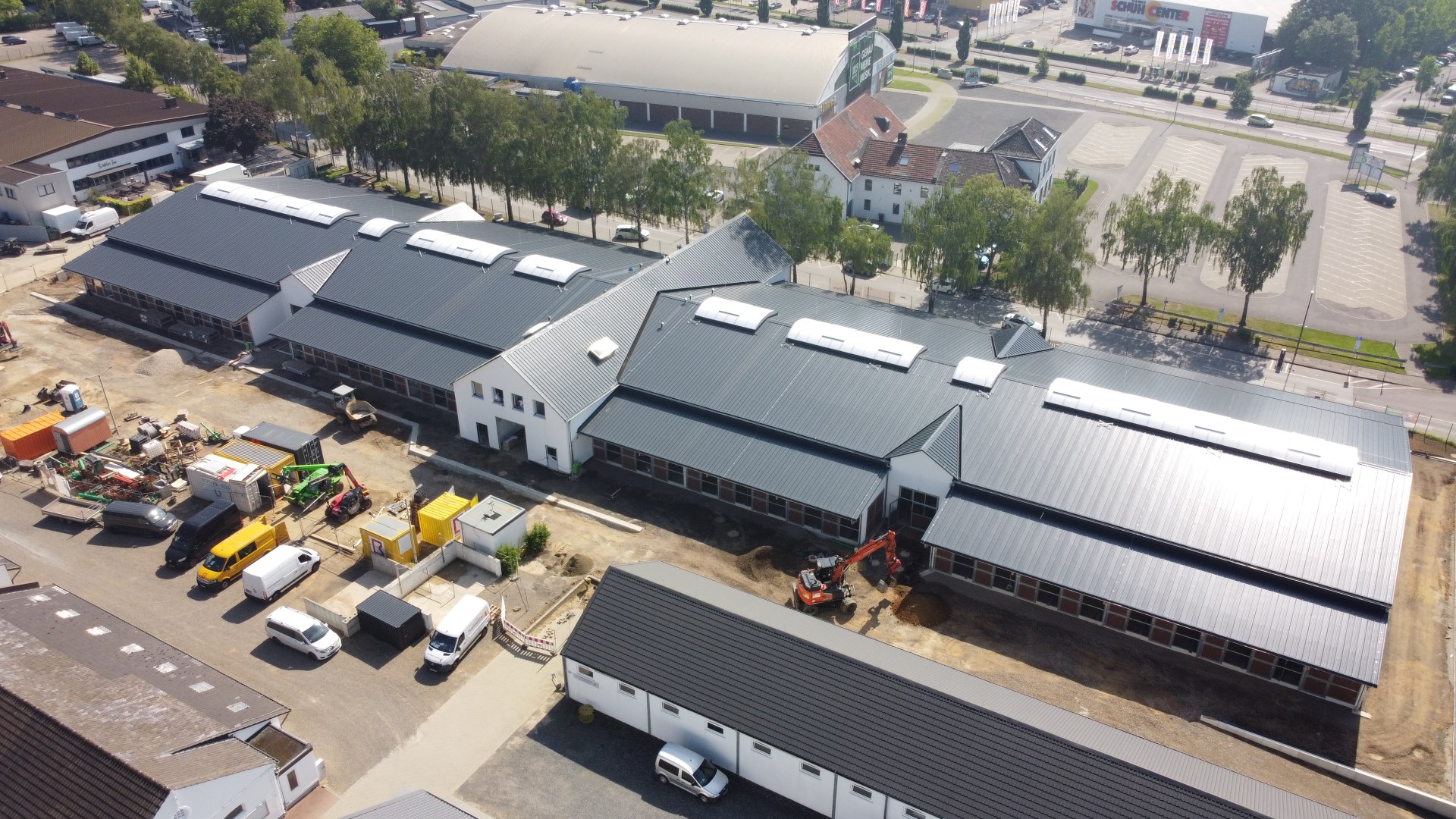 Aerial view of a newly constructed stable complex with dark roofs; construction vehicles and containers are parked in front, with trees, roads and parking areas visible in the background.