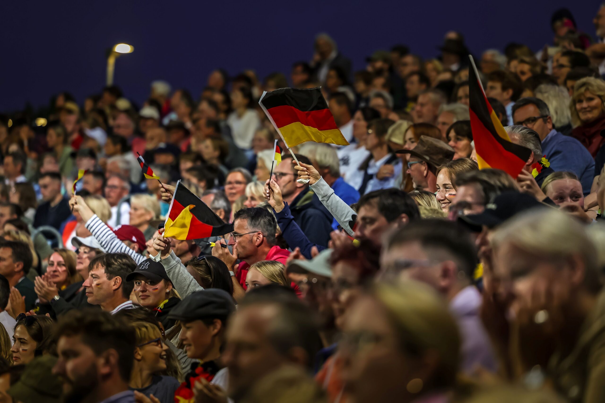 A large crowd is sitting tightly packed on the stands. Many spectators are waving German flags and applauding; the atmosphere is exuberant and festive. In the background, the sky is already dark.