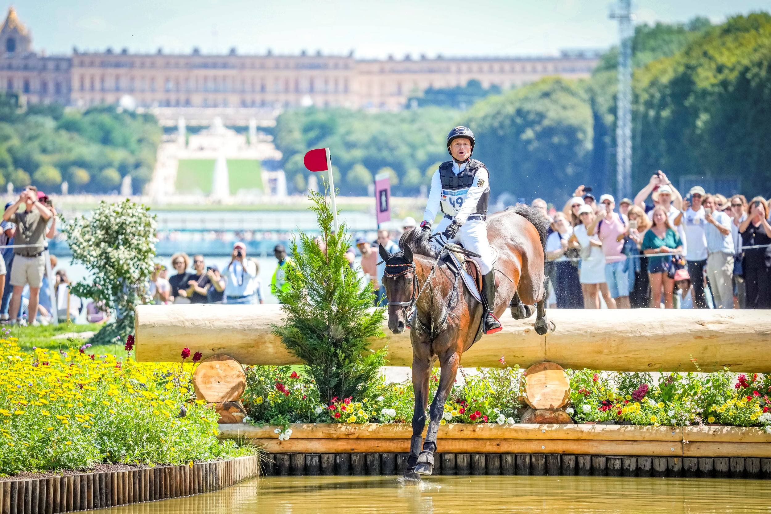 Michael Jung, dressed in white competition attire with a protective vest and start number 49, rides a brown horse over a water obstacle during the cross-country event at the Olympics in Versailles. In the background, numerous spectators stand and photograph the event, behind them is a large park with a magnificent castle. The scene is characterised by summer weather and blooming flowers.