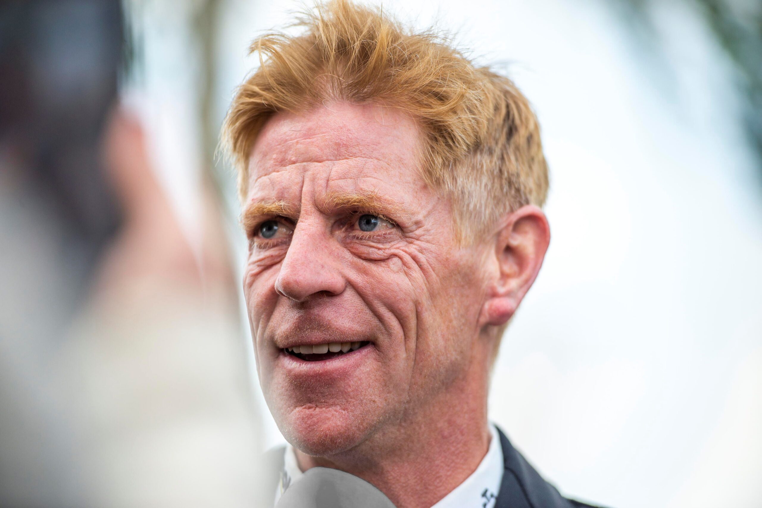 Portrait photo of show jumper Marcus Ehning with reddish-blond hair in formal riding attire, speaking into a microphone. The background is blurred, suggesting an outdoor event.