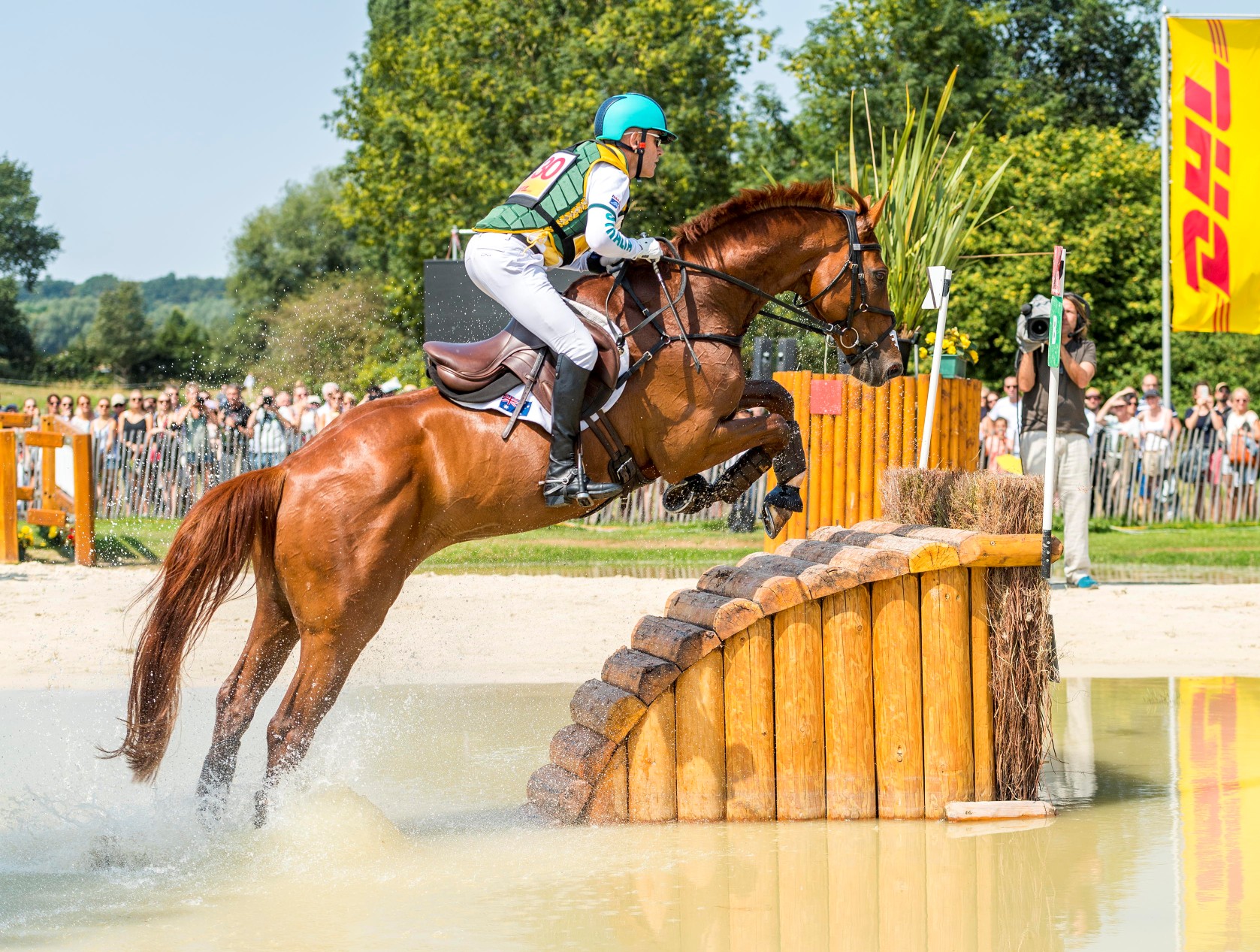 An eventing rider jumps over a solid wooden obstacle in a water jump with his chestnut horse; water splashes up; in the background, numerous spectators and a cameraman watch the cross-country course; on the right, a yellow DHL flag flutters in the wind.