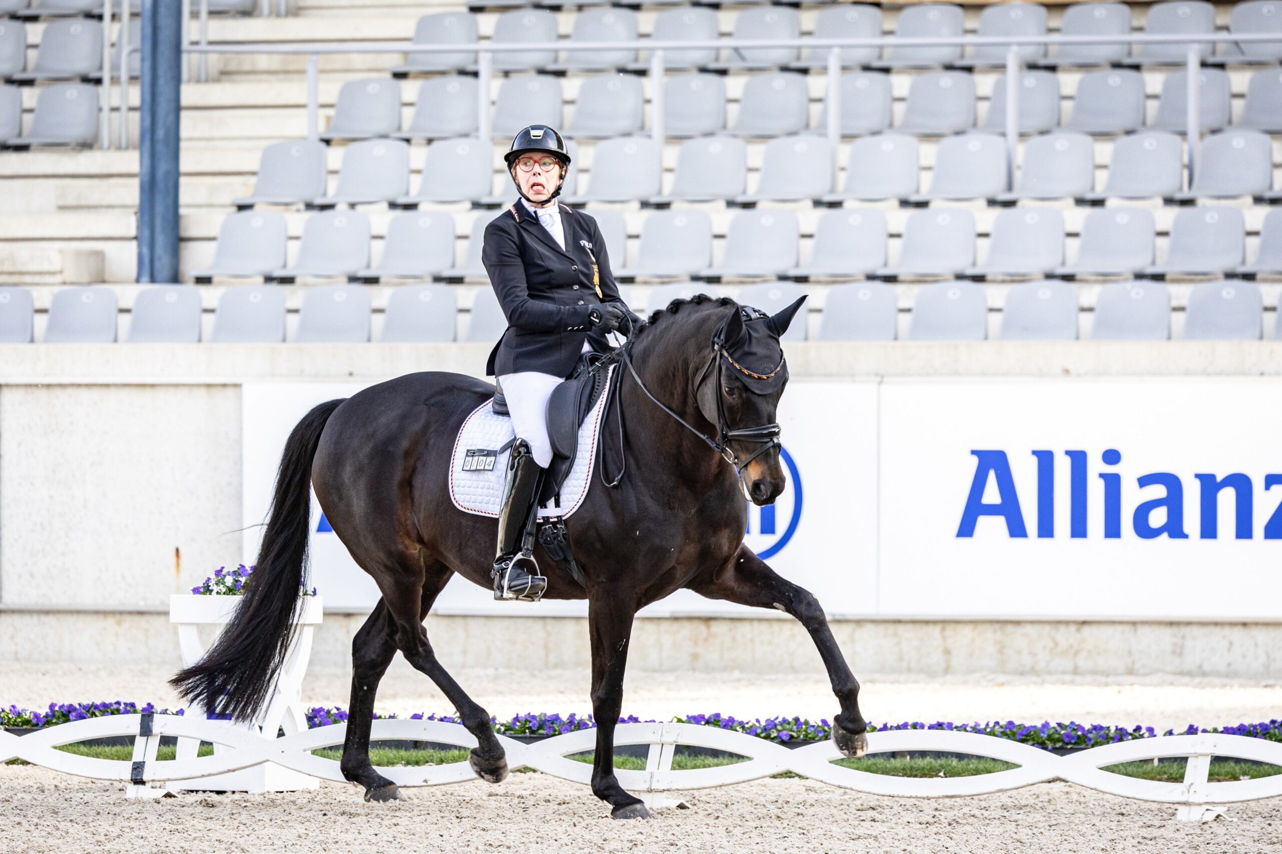 Para dressage rider in a black jacket and helmet on a dark horse performing an extended trot in a sandy arena, with grey grandstand seats and an Allianz advertising banner in the background.