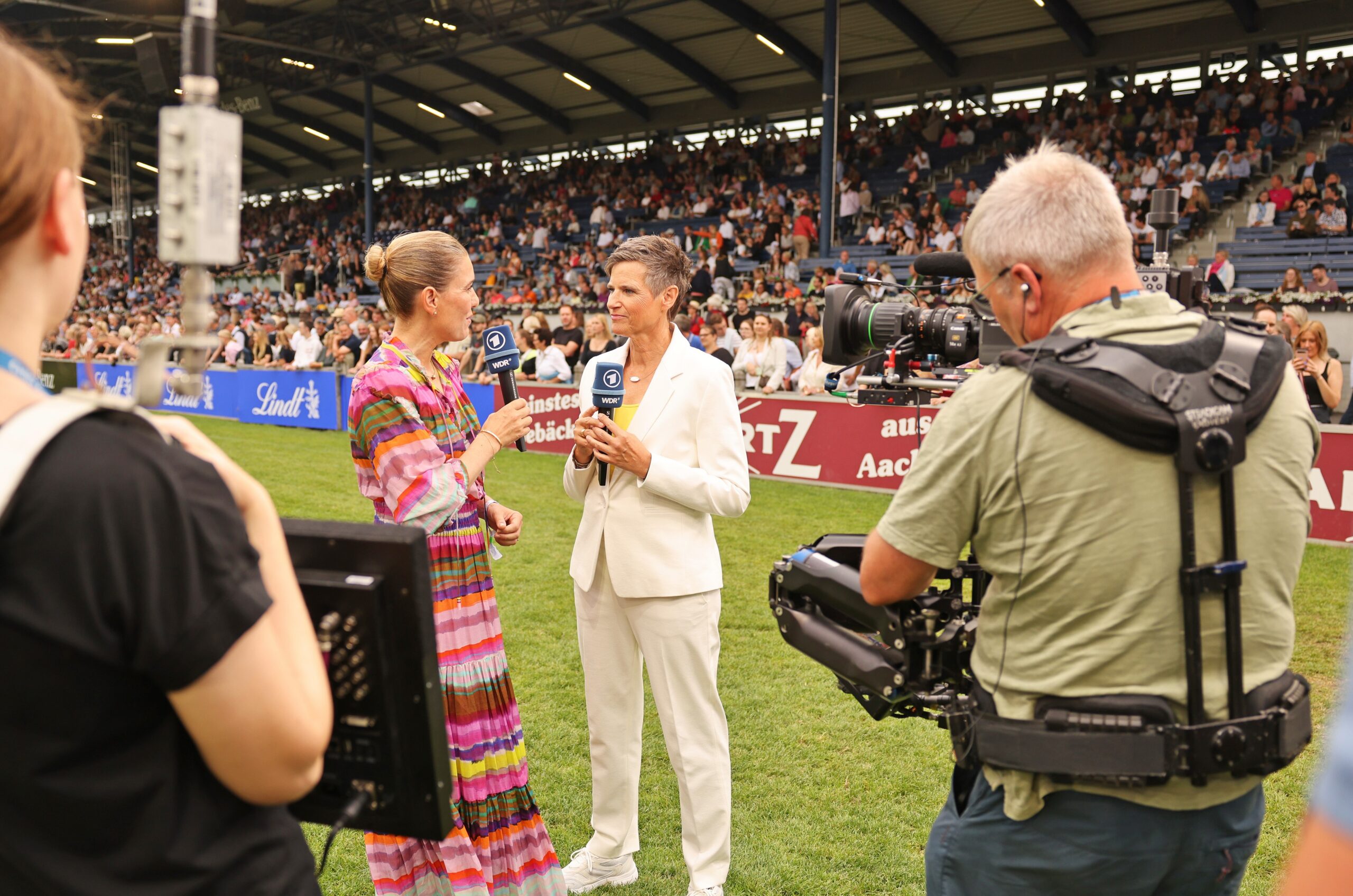 Zwei TV-Moderatorinnen stehen auf dem Rasen eines voll besetzten Reitstadions und führen ein Interview, während im Vordergrund ein Kameramann mit Steadicam und eine Assistentin mit Monitor filmen; im Hintergrund sind Zuschauertribünen und Werbebanden zu sehen.