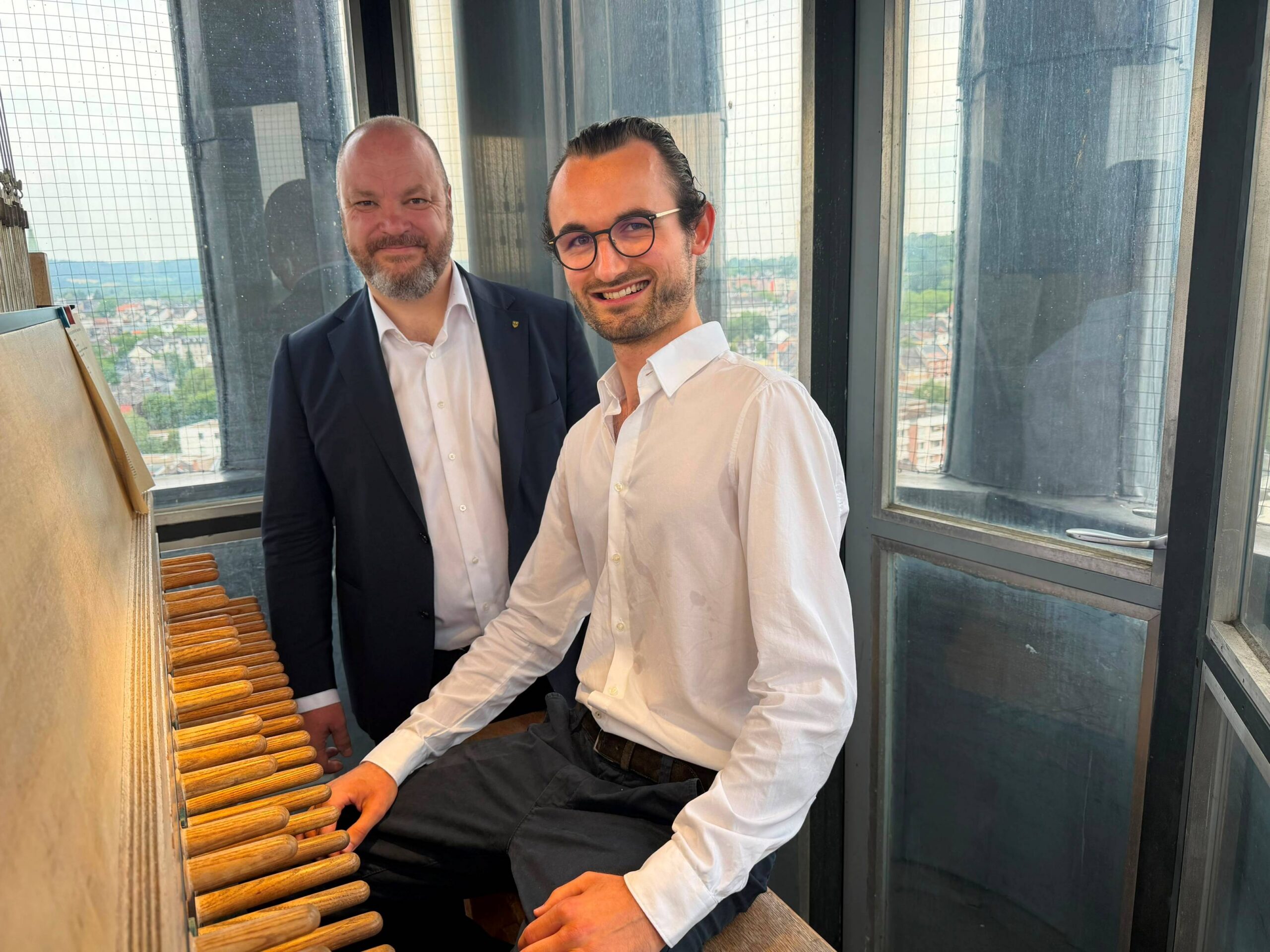 A young man in a white shirt is sitting and smiling at a carillon with many wooden levers, with a man in a dark suit standing next to him; through the surrounding windows, the cityscape of Aachen can be seen in the background.