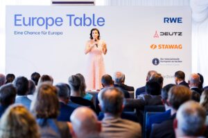 A female speaker in a pink dress is speaking into a microphone on the stage of the event ‘Europe Table – Eine Chance für Europa’ in front of a seated audience; on the right side of the stage backdrop, the logos of RWE, Deutz, STAWAG and the International Charlemagne Prize of Aachen can be seen.